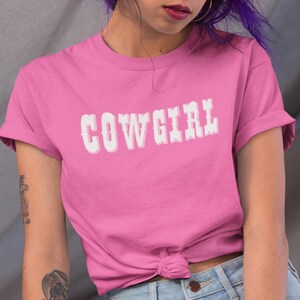 May include: A pink t-shirt featuring the word "COWGIRL" in white, decorative lettering. The shirt has short sleeves and a knot detail at the waist. The style is Western-themed.