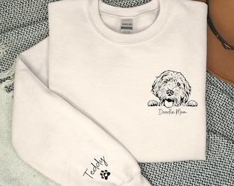 May include: White sweatshirt with a black doodle dog peeking out from the pocket and the text "Doodle Mom" underneath. The left sleeve has the text "Teddy" and a paw print.