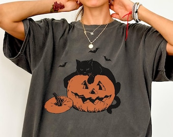 May include: Dark gray oversized t-shirt featuring a Halloween design. The graphic includes a black cat resting on a carved orange pumpkin, with bats flying above. A smaller pumpkin is next to the larger one. The word "PEPPER" is at the bottom left.