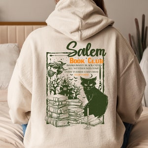 Puede incluir: Una sudadera con capucha beige con un estampado gráfico de un gato negro, una pila de libros y una casa encantada. El gráfico también incluye el texto "Salem Book Club", "Books Booze Black Cats", "All Witches Welcome", "Every Tuesday & Saturday", y "In October".