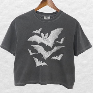 May include: Dark grey crop top with a white illustration of multiple bats in flight. The bats are of varying sizes and arranged in a dynamic composition. The shirt is hanging on a wooden hanger.