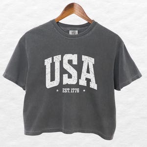 Pu&ograve; includere: Una maglietta corta grigio antracite con la scritta "USA" in lettere bianche invecchiate. Sotto la parola "USA" c'&egrave; il testo "EST. 1776" con una stella su ogni lato. La maglietta &egrave; appesa a una gruccia di legno.