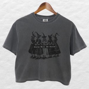 Könnte beinhalten: Dunkelgraues T-Shirt mit Rundhalsausschnitt und kurzen Ärmeln. Auf der Vorderseite befindet sich eine Schwarz-Weiß-Grafik von fünf Figuren mit Hexenhüten und dem Text "Girls Will Be Girls" darunter. Das T-Shirt hängt an einem Holzkleiderbügel.