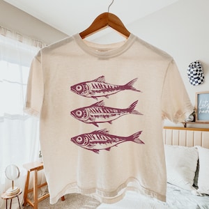 Puede incluir: Una camiseta recortada de color crema con un gr&aacute;fico de tres peces rojos en la parte delantera.
