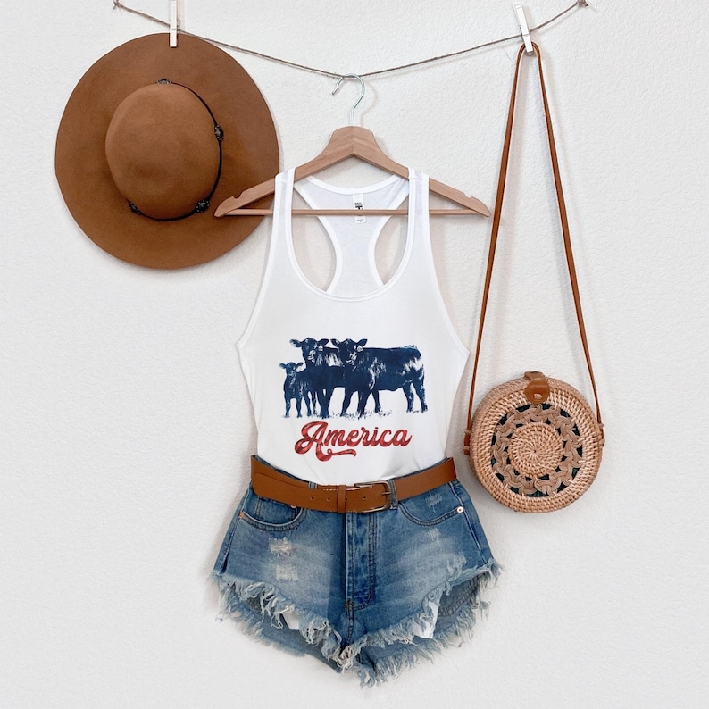 May include: White tank top with a graphic of cows and the text "America" in red and blue. The tank top is paired with denim shorts and a brown woven bag.