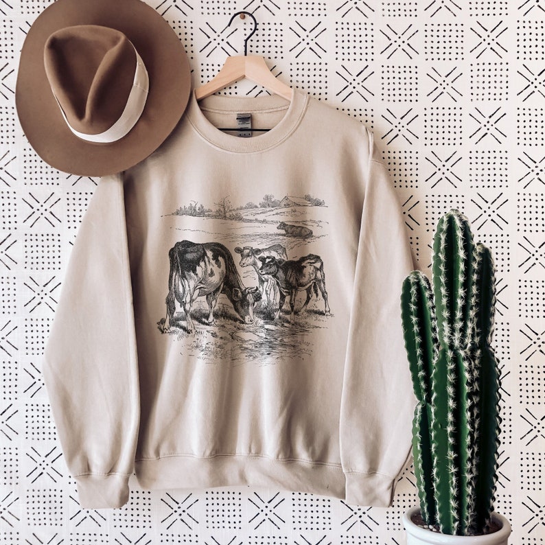 K&ouml;nnte beinhalten: Ein beiger Pullover mit einer Schwarz-Wei&szlig;-Illustration von K&uuml;hen auf einer Weide. Der Pullover hat einen Rundhalsausschnitt und lange &Auml;rmel.