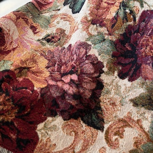 May include: Close-up of a woven throw blanket featuring a floral pattern in shades of burgundy, gold, and green. The design includes large, detailed flowers and scrolling accents against a cream background. The blanket has a fringed edge.