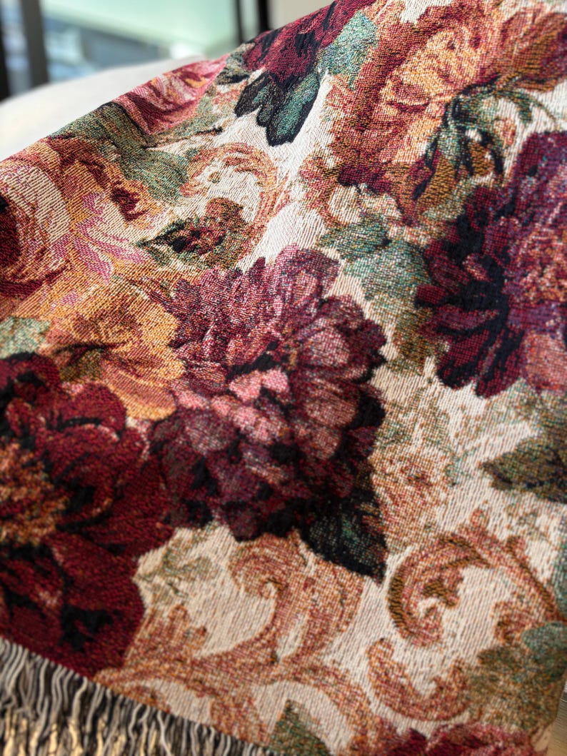 May include: Close-up of a woven throw blanket featuring a floral pattern in shades of burgundy, gold, and green. The design includes large, detailed flowers and scrolling accents against a cream background. The blanket has a fringed edge.