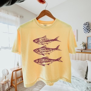 Puede incluir: Una camiseta amarilla con un gr&aacute;fico de tres peces estilizados en marr&oacute;n. Los peces est&aacute;n dispuestos en una pila vertical, con el pez m&aacute;s grande en la parte superior y el pez m&aacute;s peque&ntilde;o en la parte inferior.