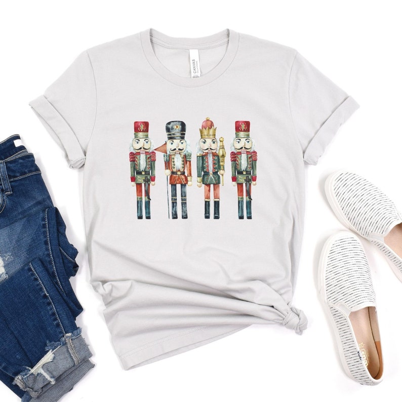 May include: A light gray t-shirt featuring a watercolor illustration of four toy nutcrackers. The nutcrackers are in various colors, including red, green, and gold. The shirt is paired with blue denim shorts and white slip-on shoes.