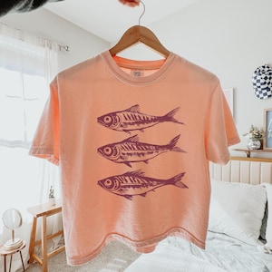 Puede incluir: Una camiseta recortada de color melocot&oacute;n con un gr&aacute;fico de tres peces estilizados en color rosa oscuro. Los peces est&aacute;n dispuestos verticalmente, con el pez m&aacute;s grande en la parte superior y el pez m&aacute;s peque&ntilde;o en la parte inferior.