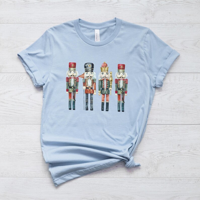 May include: Light blue t-shirt featuring a watercolor illustration of four toy nutcrackers. The nutcrackers are dressed in red, green, and black uniforms with gold accents. The shirt is made of soft material.