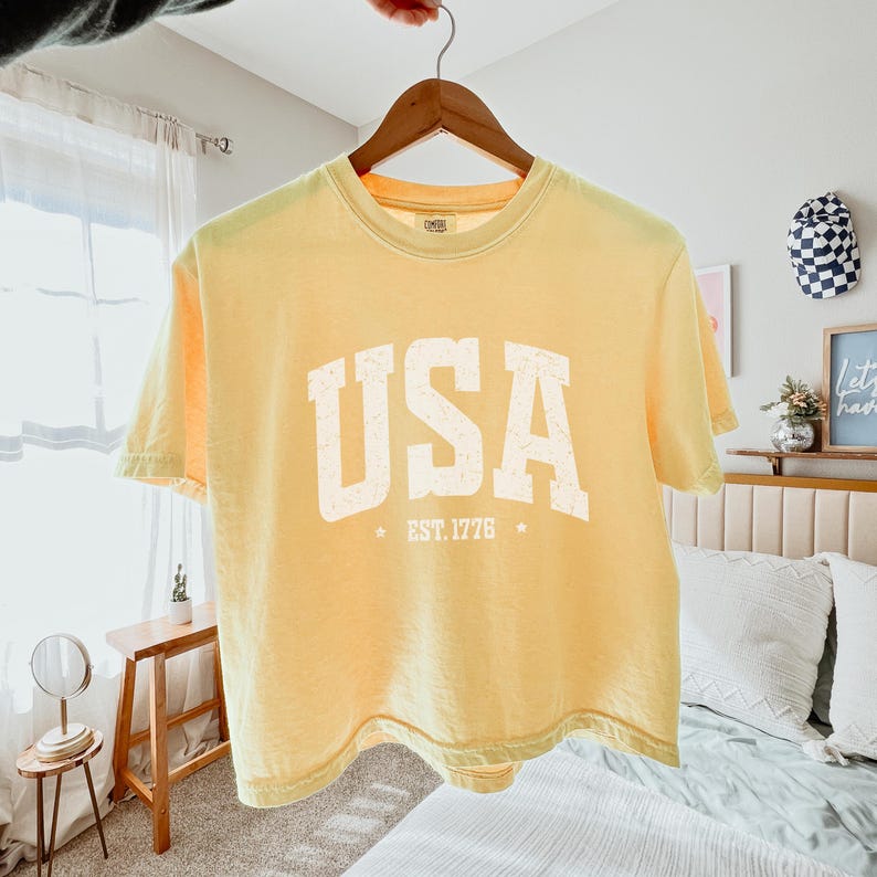 Pu&ograve; includere: Una maglietta giallo chiaro con la scritta "USA" in grandi lettere bianche sbiadite. Sotto c'&egrave; il testo "EST. 1776" con stelle. La maglietta &egrave; appesa a una gruccia di legno.