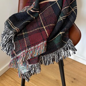 May include: A woven plaid throw blanket in shades of black, red, green, and cream, draped over a brown leather chair. The blanket features a fringed edge with a mix of colorful threads. The pattern is a classic tartan design.