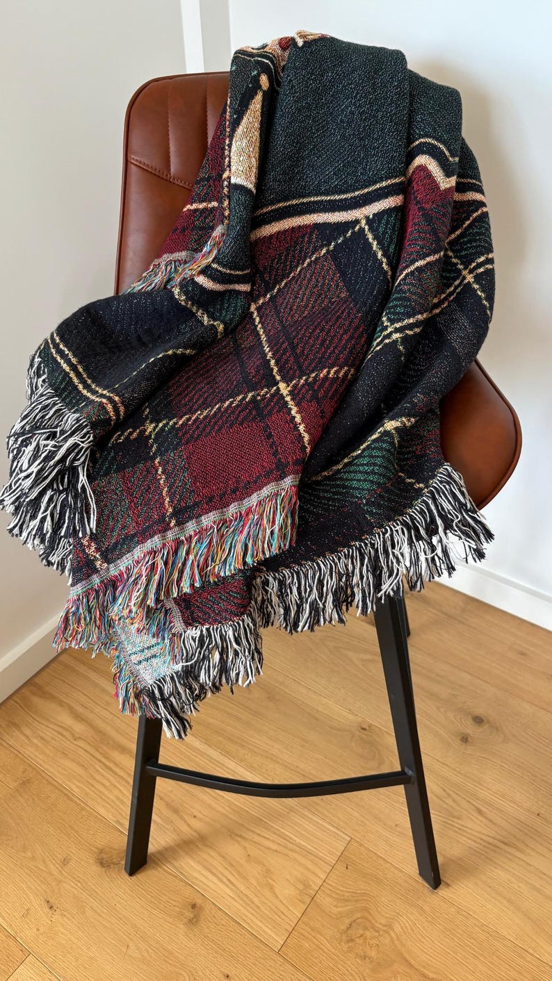 May include: A woven plaid throw blanket in shades of black, red, green, and cream, draped over a brown leather chair. The blanket features a fringed edge with a mix of colorful threads. The pattern is a classic tartan design.