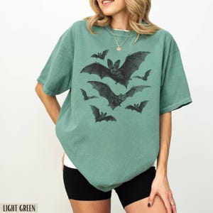 May include: A light green t-shirt with a graphic print of bats in black. The shirt has a relaxed fit and short sleeves.