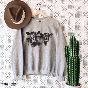 K&ouml;nnte beinhalten: Sportgraues Sweatshirt mit einer schwarz-wei&szlig;en Illustration von zwei K&uuml;hen. Das Sweatshirt h&auml;ngt an einem Holzkleiderb&uuml;gel. Ein brauner Hut mit wei&szlig;em Band befindet sich links. Rechts befindet sich ein Kaktus. Der Text "SPORT GREY" ist unten.