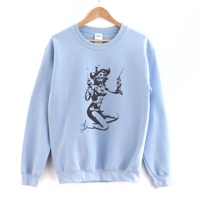 Pu&ograve; includere: Felpa blu chiaro con una stampa grafica in bianco e nero di una donna con un cappello da cowboy e che tiene due pistole.