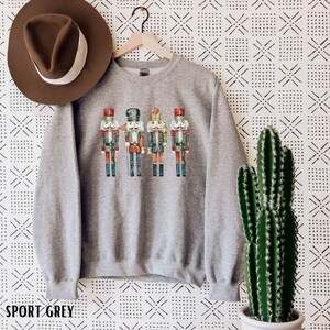 May include: A heather gray sweatshirt featuring a colorful illustration of four nutcrackers in festive uniforms. The text "SPORT GREY" is printed at the bottom. A brown hat hangs on the left side of the sweatshirt.