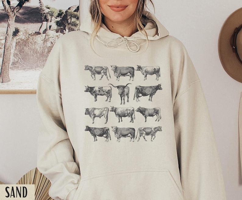 May include: A cream-colored hoodie with a graphic print of 12 black and white cows. The cows are arranged in four rows of three.