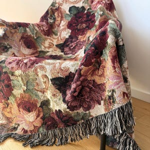 May include: A floral tapestry throw blanket draped over a chair. The blanket features a pattern of large, colorful roses in shades of burgundy, peach, and cream, with a black and white fringe.