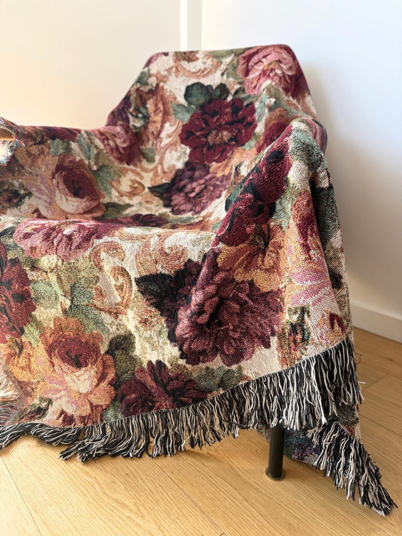 May include: A floral tapestry throw blanket draped over a chair. The blanket features a pattern of large, colorful roses in shades of burgundy, peach, and cream, with a black and white fringe.