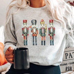 May include: A light gray sweatshirt featuring a watercolor illustration of four toy nutcracker soldiers. The nutcrackers are in red, green, and brown uniforms with gold accents. The word "ASH" is printed on the bottom right.