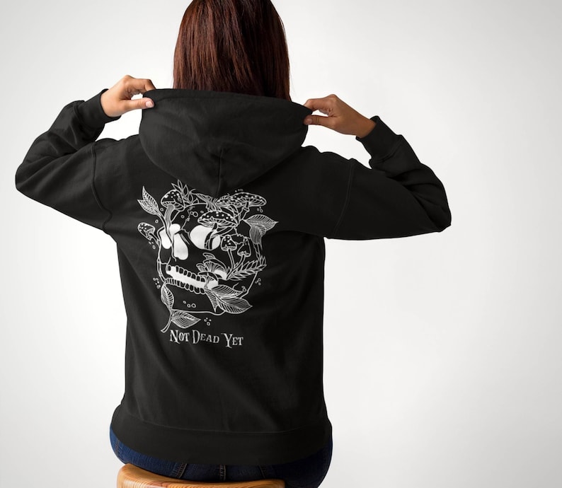 May include: Black hooded sweatshirt with a white graphic on the back. The graphic features a skull surrounded by mushrooms and foliage, with the text "NOT DEAD YET" below. The person is wearing blue jeans.