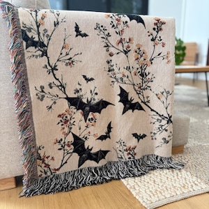 May include: Beige throw blanket with a black bat and floral pattern. The blanket has a fringed edge with a mix of colors. The design features black bats and branches with orange and red flowers.