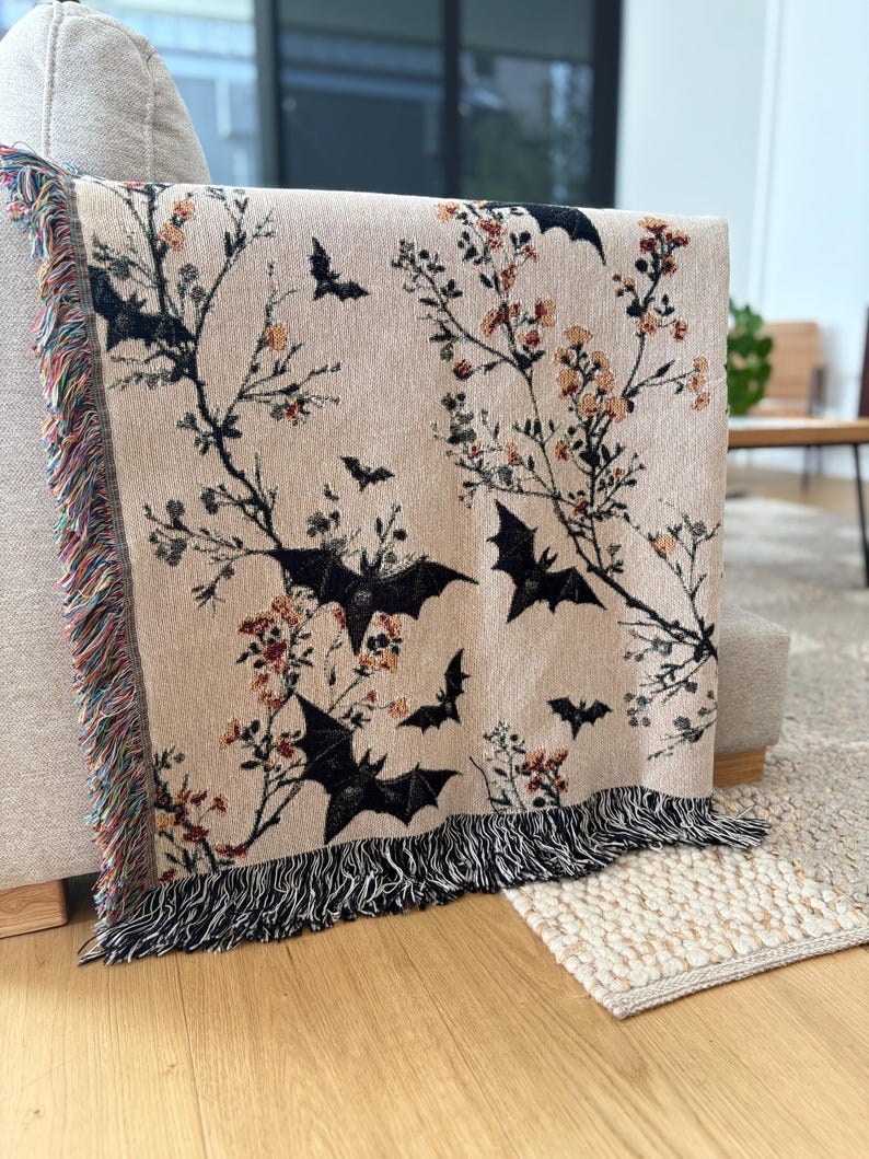 May include: Beige throw blanket with a black bat and floral pattern. The blanket has a fringed edge with a mix of colors. The design features black bats and branches with orange and red flowers.