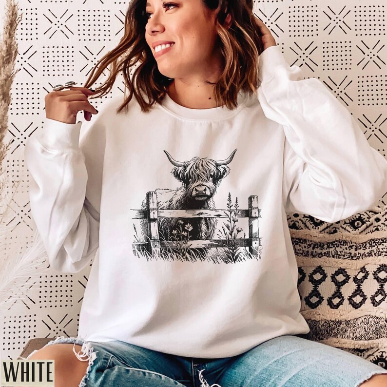 Puede incluir: Sudadera blanca con una ilustraci&oacute;n en blanco y negro de una vaca de las tierras altas de Escocia de pie detr&aacute;s de una valla con flores.
