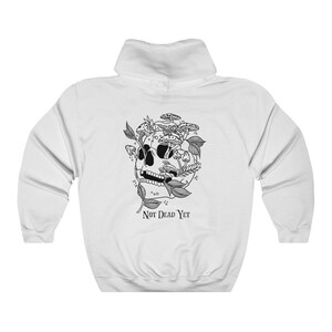 May include: White hooded sweatshirt with a black graphic of a skull with mushrooms and foliage. The text "NOT DEAD YET" is below the skull. The sweatshirt has a drawstring hood and a kangaroo pocket.