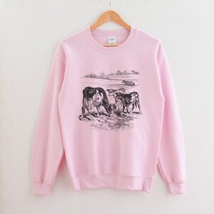 K&ouml;nnte beinhalten: Ein rosa Sweatshirt mit einer Schwarzwei&szlig;illustration von K&uuml;hen auf einer Weide. Die K&uuml;he stehen sich gegen&uuml;ber und scheinen zu grasen.