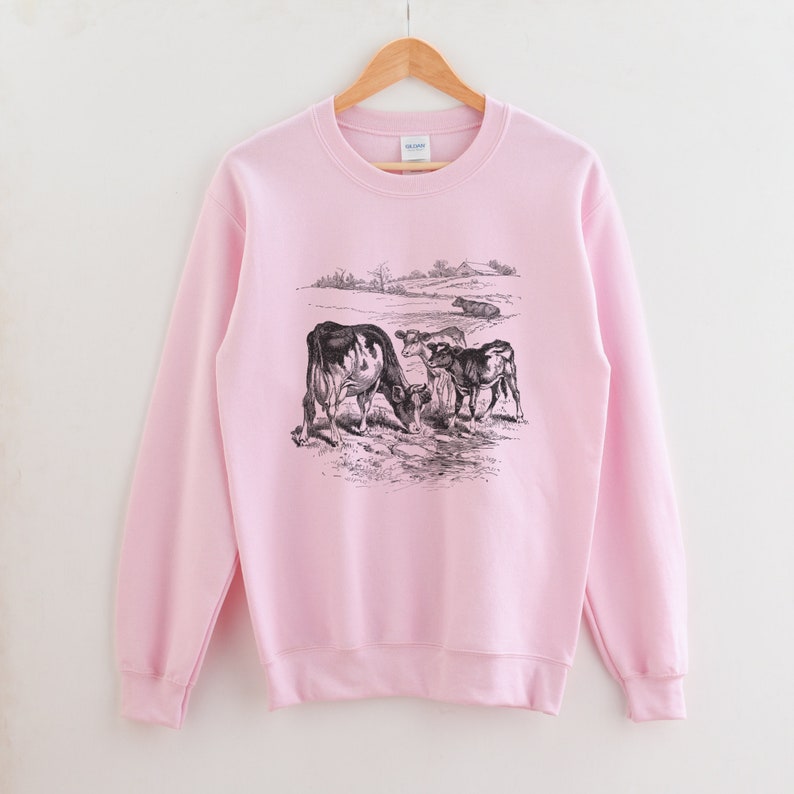 K&ouml;nnte beinhalten: Ein rosa Sweatshirt mit einer Schwarzwei&szlig;illustration von K&uuml;hen auf einer Weide. Die K&uuml;he stehen sich gegen&uuml;ber und scheinen zu grasen.