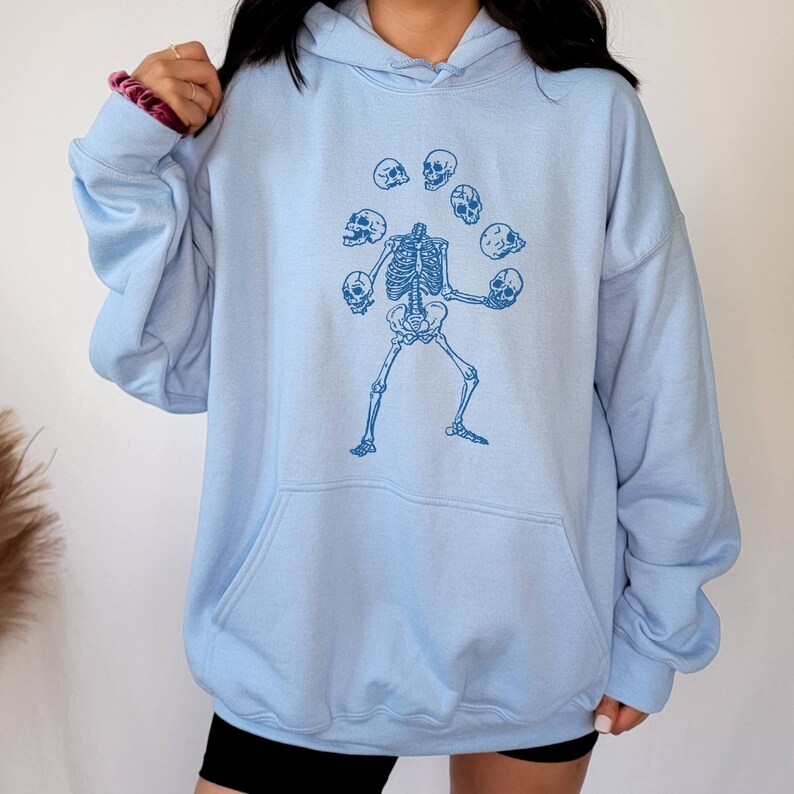 May include: A light blue hoodie featuring a graphic of a skeleton juggling skulls. The skeleton is wearing a blue shirt and has a blue tie. The skulls are all different colours.