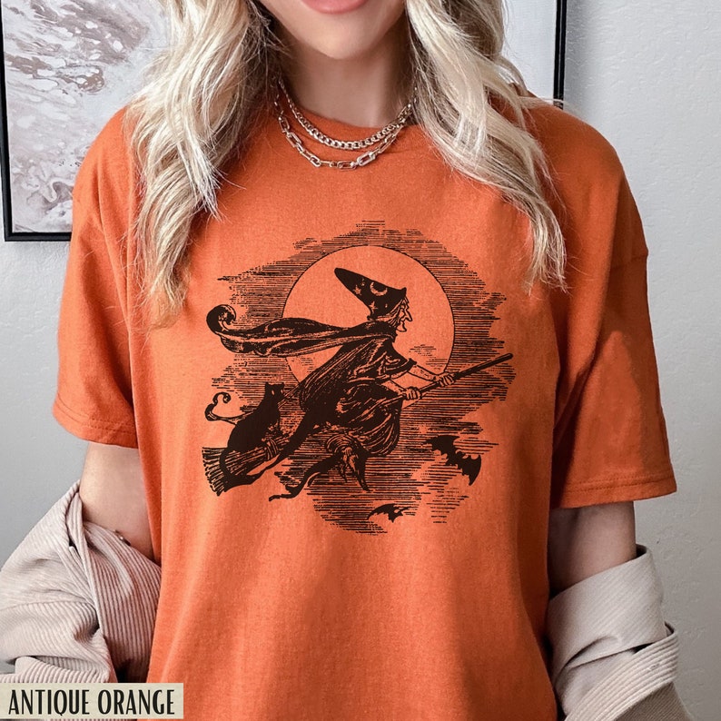 May include: Antique orange t-shirt featuring a black illustration of a witch on a broomstick with a black cat and bats, set against a full moon. The text "ANTIQUE ORANGE" is at the bottom of the shirt.