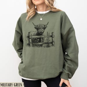 Puede incluir: Una sudadera verde con una ilustraci&oacute;n en blanco y negro de una vaca de las tierras altas de Escocia de pie detr&aacute;s de una valla. La vaca est&aacute; mirando hacia la izquierda de la imagen. El texto "MILITARY GREEN" est&aacute; en la parte inferior de la imagen.
