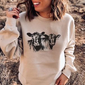 K&ouml;nnte beinhalten: Beiges Sweatshirt mit Rundhalsausschnitt und einer schwarz-wei&szlig;en Illustration von zwei K&uuml;hen. Die K&uuml;he haben Ohrmarken. Das Sweatshirt hat lange &Auml;rmel und eine lockere Passform. Das Design ist mittig auf der Vorderseite platziert.