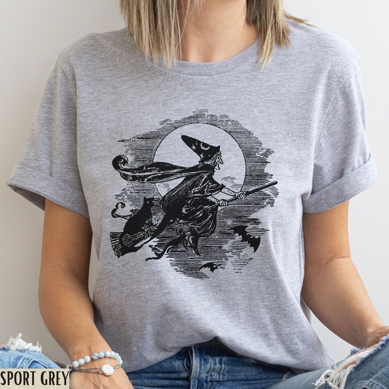May include: Sport grey t-shirt featuring a black and white illustration of a witch riding a broom in front of a full moon, with a black cat and bats. The shirt has short sleeves and a crew neck.