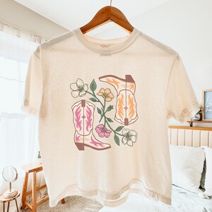 May include: A cream-coloured t-shirt with a graphic of two cowboy boots in pink and orange, with floral accents. The boots are set against a backdrop of flowers and leaves. The shirt is hanging on a wooden hanger.