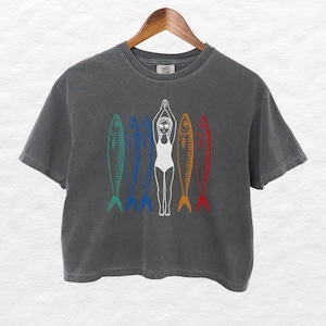 以下が含まれることがあります： 濃いグレーのクロップドTシャツに、水着を着た女性が5匹の魚の間で立っているグラフィックがプリントされています。魚は一列に並んでおり、それぞれ異なる色（青、緑、赤、オレンジ、黄色）で縁取られています。