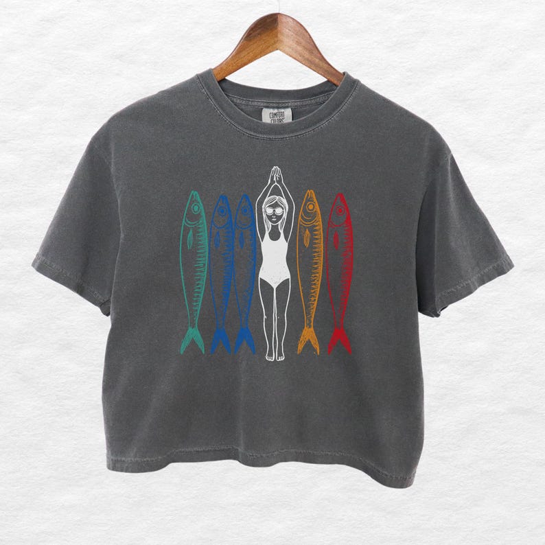 以下が含まれることがあります： 濃いグレーのクロップドTシャツに、水着を着た女性が5匹の魚の間で立っているグラフィックがプリントされています。魚は一列に並んでおり、それぞれ異なる色（青、緑、赤、オレンジ、黄色）で縁取られています。
