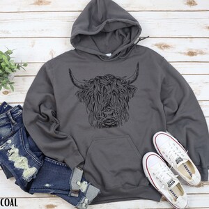 May include: A charcoal gray hooded sweatshirt with a black and white illustration of a Highland cow on the front. The sweatshirt is laying on a wooden surface with a pair of white canvas shoes and a pair of blue jeans.