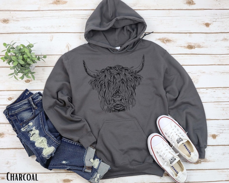 May include: A charcoal gray hooded sweatshirt with a black and white illustration of a Highland cow on the front. The sweatshirt is laying on a wooden surface with a pair of white canvas shoes and a pair of blue jeans.