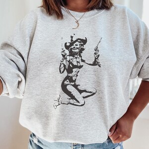Pu&ograve; includere: Una felpa grigia con una stampa grafica in bianco e nero di una donna che indossa un cappello da cowboy e tiene due pistole.