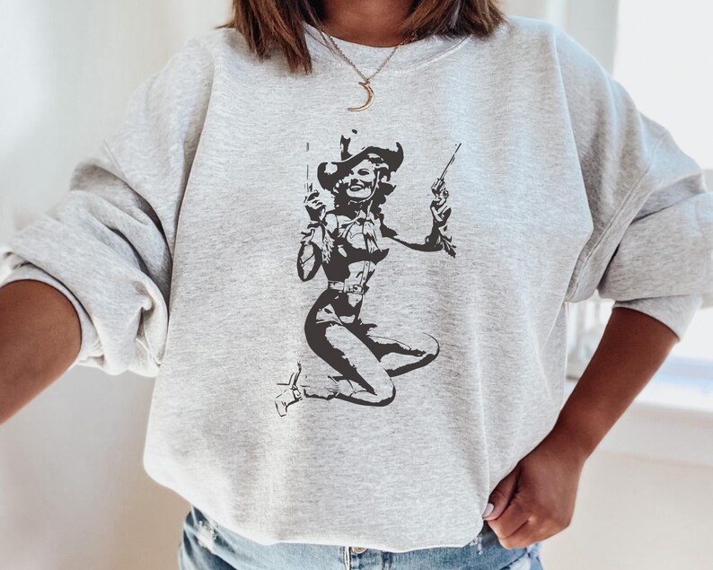 Pu&ograve; includere: Una felpa grigia con una stampa grafica in bianco e nero di una donna che indossa un cappello da cowboy e tiene due pistole.