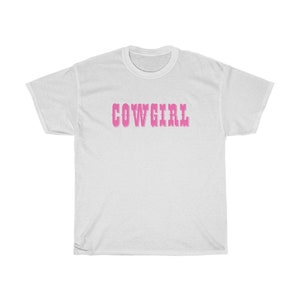 May include: White t-shirt featuring the word "COWGIRL" in pink, decorative lettering across the chest. The shirt has short sleeves and a crew neck. The design is simple and bold, suitable for casual wear.