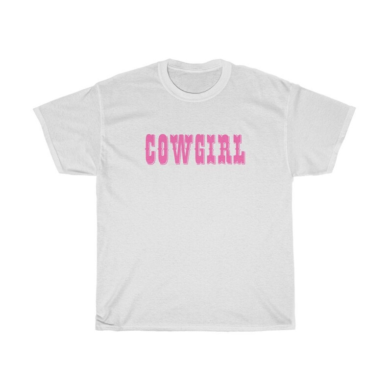 May include: White t-shirt featuring the word "COWGIRL" in pink, decorative lettering across the chest. The shirt has short sleeves and a crew neck. The design is simple and bold, suitable for casual wear.
