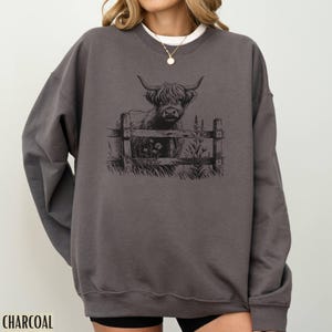 Puede incluir: Una sudadera gris antracita con una ilustraci&oacute;n en blanco y negro de una vaca de las tierras altas mirando por encima de una valla. La vaca est&aacute; de pie en un campo con hierba y flores.