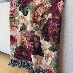 May include: A floral tapestry throw blanket with a cream background and a pattern of burgundy, pink, and green flowers. The blanket has a fringe trim with black and white threads. The blanket is draped over a light gray sofa.